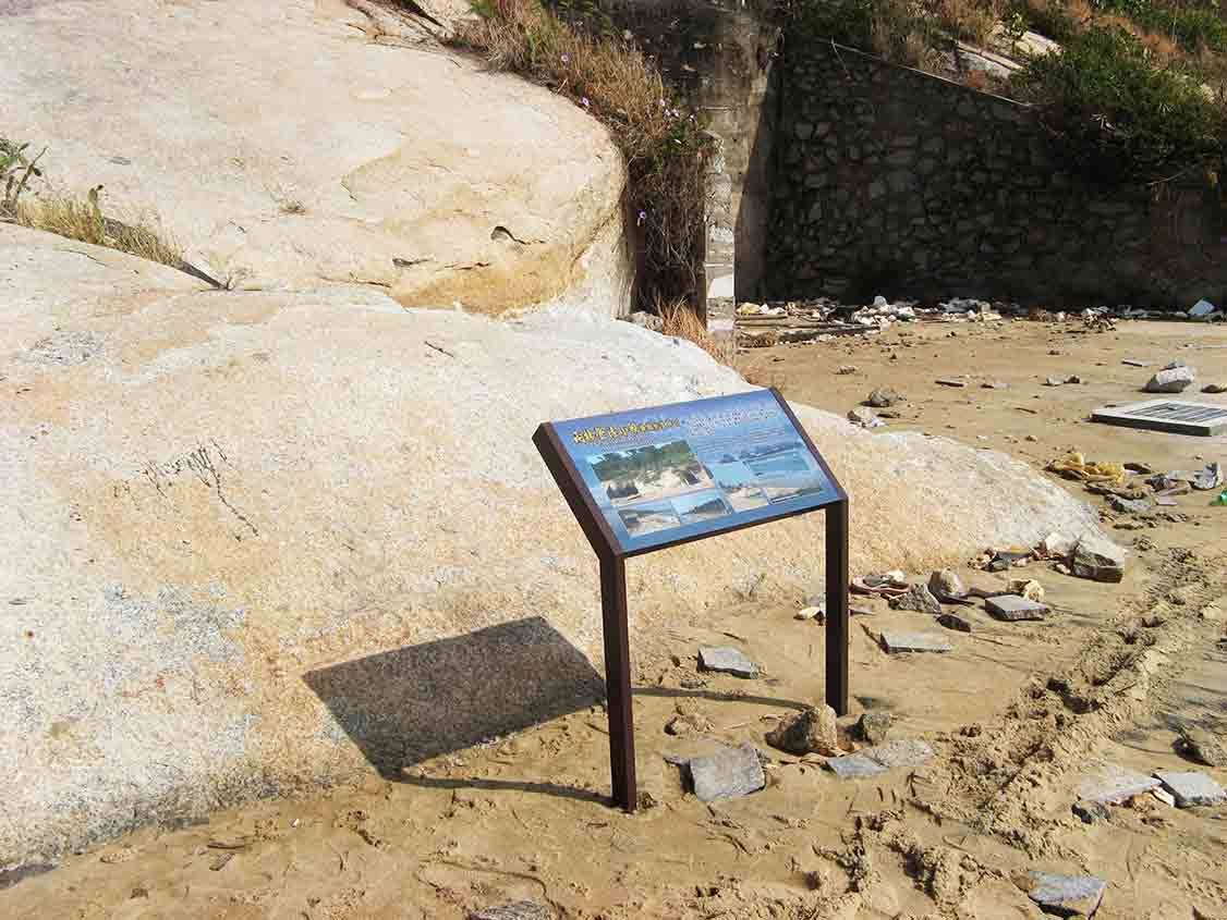 搶灘坑道藍色解說牌設於海岸沙地，背景花崗岩塊與石砌護坡延伸至沙灘