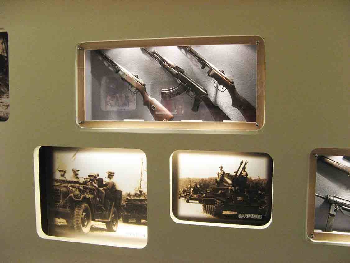 湖井頭戰史館橄欖綠牆面壓克力外掛展示框，內陳三把繳獲長槍特寫，下方圓角燈箱呈現吉普車隊與戰車隊伍泛黃舊照