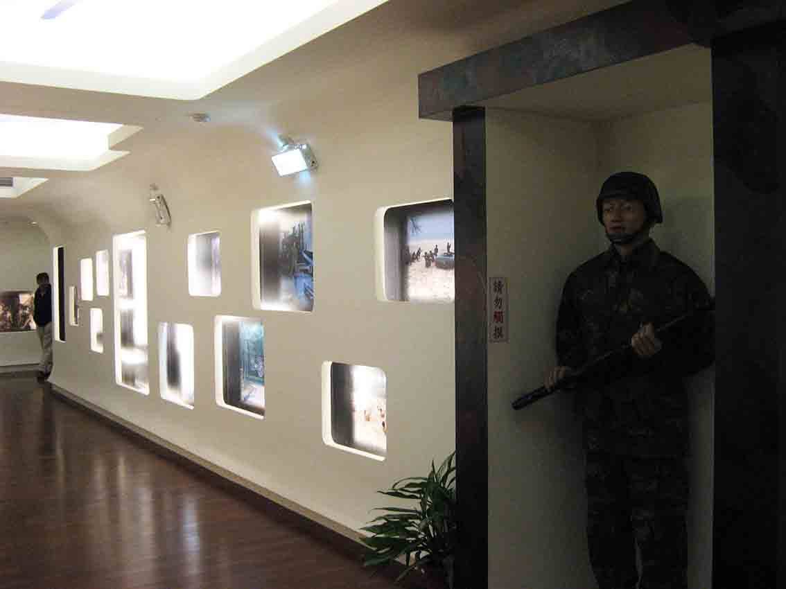 湖井頭戰史館弧形長廊展牆全景，沿牆排列大小不一的圓角燈箱戰地照片，右側深色立柱旁設持槍站哨憲兵人形立牌與「請勿觸摸」警語