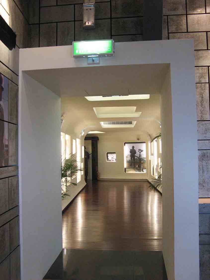 湖井頭戰史館入口框景，仿石材外牆與綠色逃生標示下方框出弧形長廊縱深，遠景可見圓角燈箱展示窗與木紋地板倒影