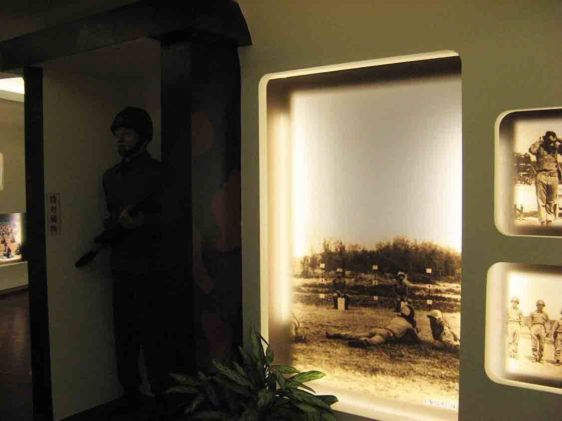 湖井頭戰史館迷彩柱與站哨國軍人形立牌旁，大型黑白燈箱呈現靶場射擊訓練場景，右側嵌牆圓角窗格陳列舊照