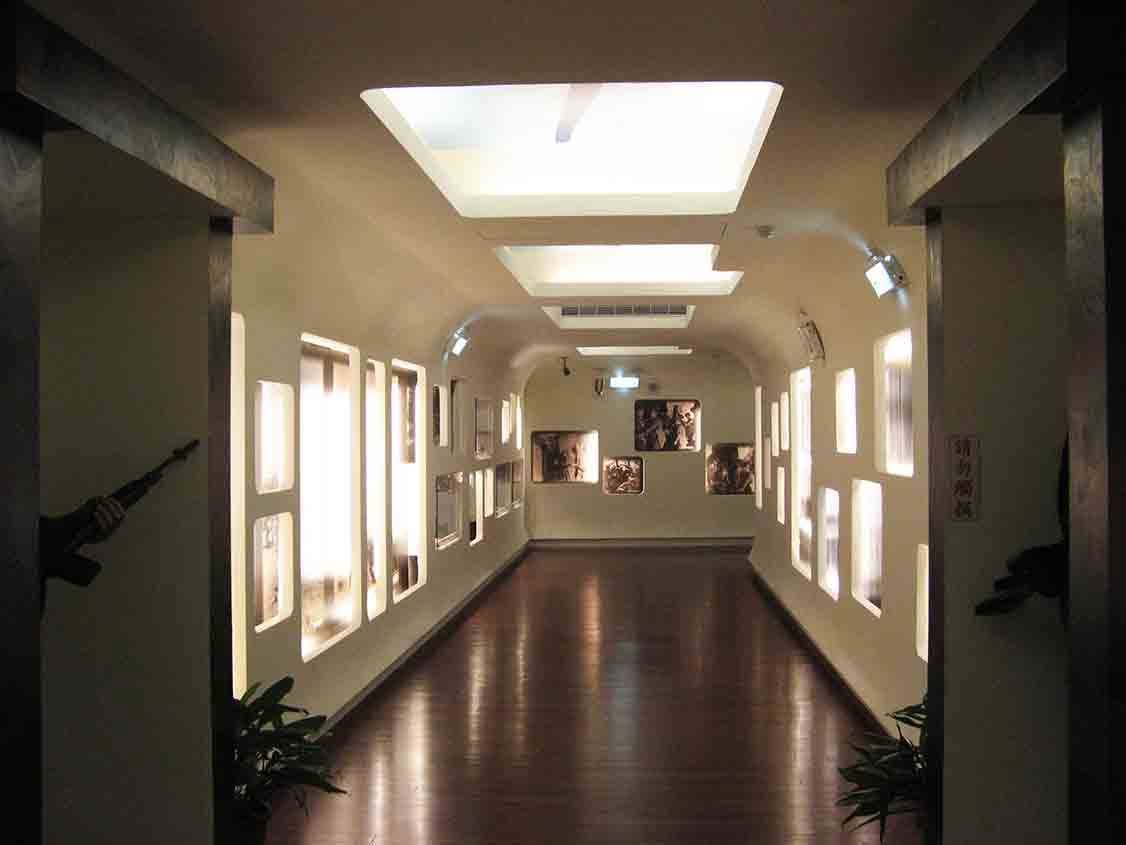 湖井頭戰史館主走廊縱深透視，木紋地板兩側延伸圓角燈箱窗格，盡頭懸掛戰地照片，兩側迷彩柱旁設站哨國軍人形立牌