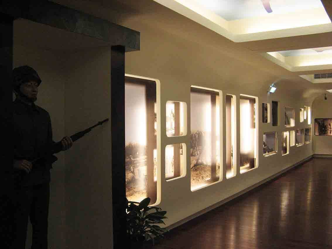 湖井頭戰史館長廊式暖光展廳全景，迷彩柱旁站哨國軍人形立牌與沿牆延伸的圓角窗格戰地照片陣列