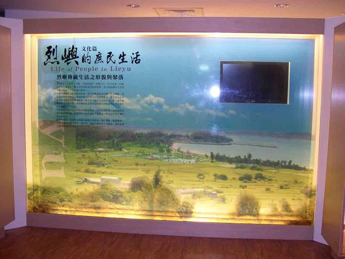 烈嶼鄉史館「烈嶼的庶民生活 Life of People in Lieyu」文化篇主視覺燈箱，藍天搭配小金門海岸聚落空拍與標題書法