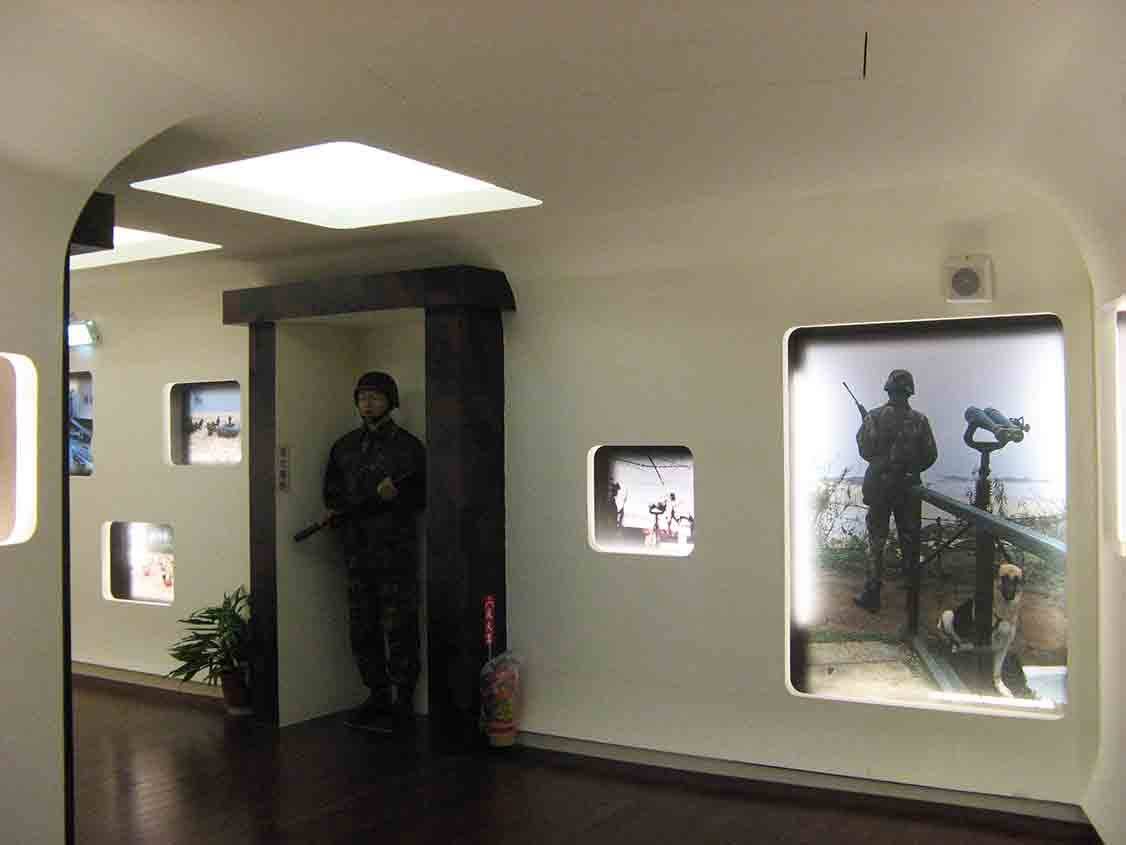 湖井頭戰史館仿坑道入口場景，門邊豎立持槍站哨國軍人形立牌，右側大圖呈現觀測哨兵與軍犬剪影