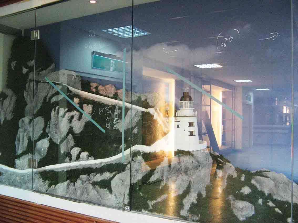 基隆碼頭展示窗內的馬祖燈塔立體模型，玻璃牆內陳列岩礁地形與東引白色燈塔，玻璃面上仍留有施工標記