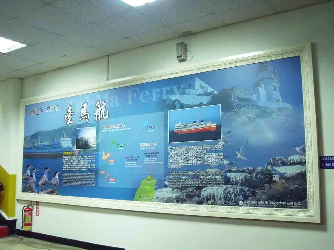 基隆碼頭大廳橫幅大圖「臺馬航 Tai-Ma Ferry」，藍色海面背景結合燈塔、輪船與航線地圖，外框以白色雕花鏡框裝飾