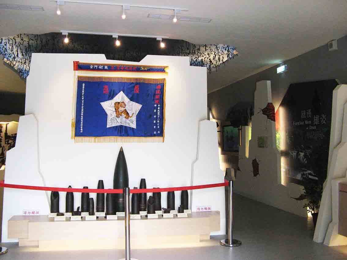 湖井頭戰史館入口「金門忠貞部隊」虎頭錦旗與下方陳列各式砲彈彈頭，右側通往「鐵漢雄姿 Forging Men of Iron」展區