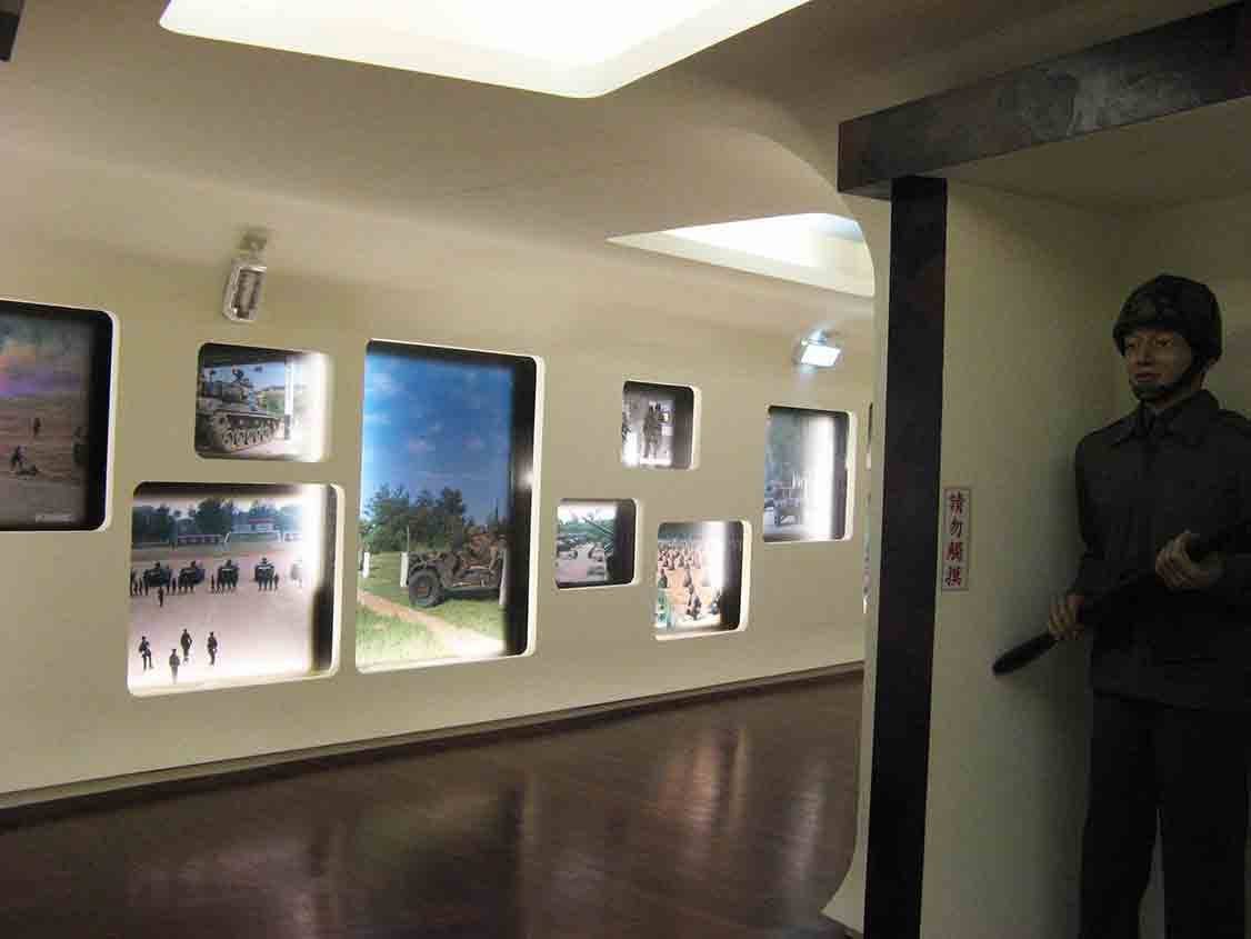 湖井頭戰史館長廊式展牆，多幅戰地操演與裝甲車照片嵌入圓角窗格，盡頭立有持槍國軍人形立牌