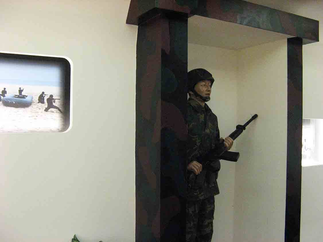 湖井頭戰史館迷彩立柱旁站哨國軍人形立牌特寫，左側嵌牆圓角窗格內呈現橡皮艇海上突擊訓練彩色照片