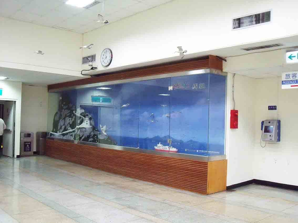 基隆碼頭候船大廳「來去馬祖」整面玻璃展示牆，藍色海洋背景搭配燈塔、海鳥、輪船剪影，下方木格柵基座與時鐘指示