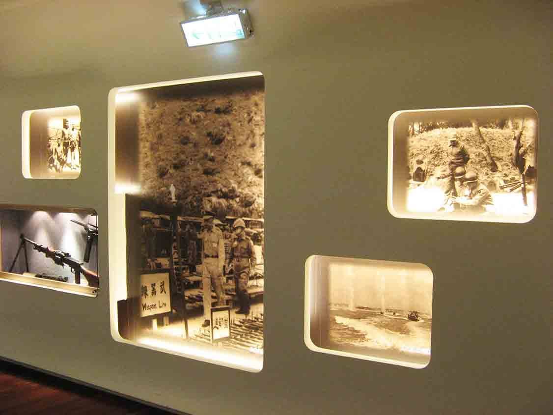 湖井頭戰史館「武器陳列 Weapon Line」主題展牆全景，中央大圖呈現部隊整裝列隊，周圍嵌入坑道作戰、海灘登陸與機槍實體展示