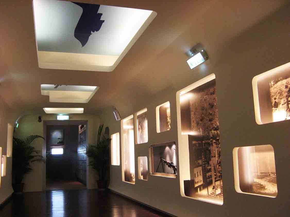 湖井頭戰史館弧形長廊透視，米白天花板嵌入金門精神堡壘剪影燈箱，右側牆面延伸圓角燈箱戰地大圖，盡頭通往坑道防火門