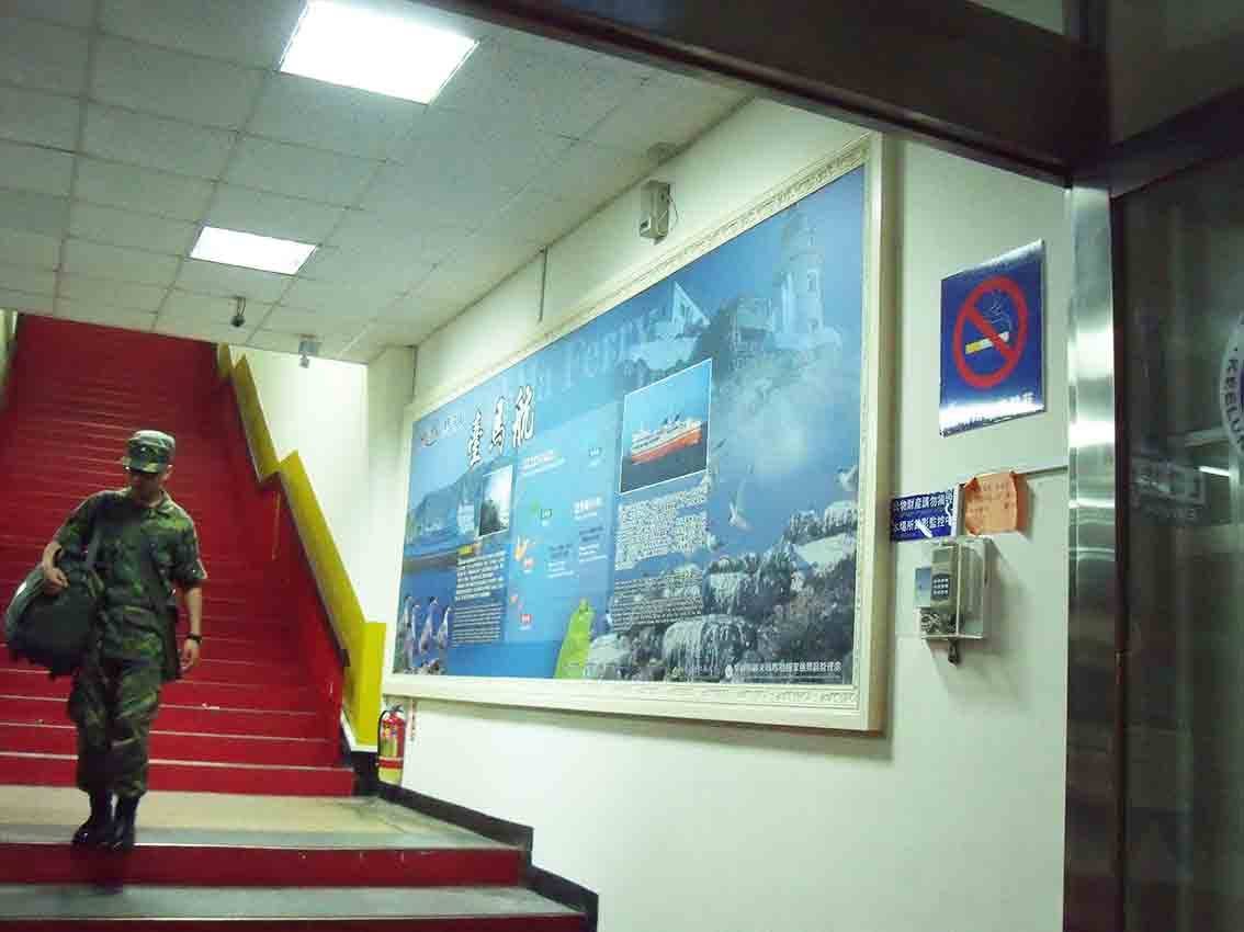 基隆碼頭候船大廳樓梯旁懸掛「臺馬航 Tai-Ma Ferry」大圖框架，藍色海景中嵌入燈塔與輪船影像，紅色階梯上一名軍人正走下