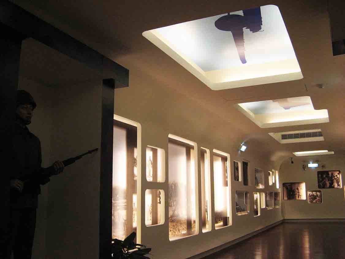 湖井頭戰史館主走廊仰視，米白天花板嵌入金門精神堡壘剪影燈箱，左側站哨國軍人形立牌與沿牆延伸的圓角燈箱展示窗