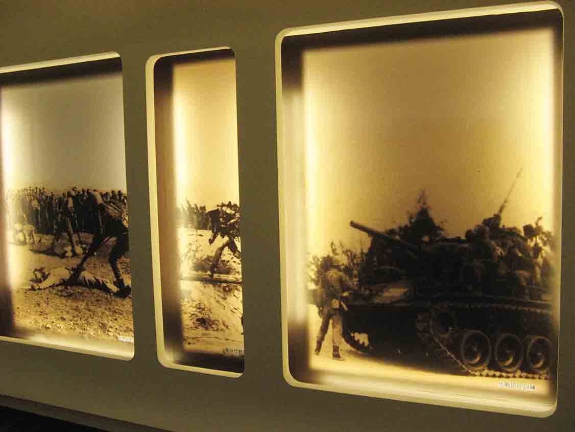 湖井頭戰史館嵌牆圓角燈箱展示牆，三幅泛黃黑白歷史照片呈現衝鋒部隊、戰場行軍與戰車載運兵員的戰役場景