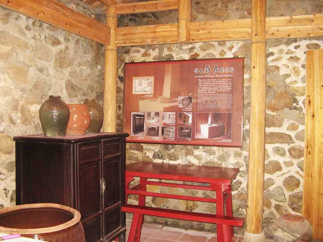 馬祖后里文物館仿古廚房場景轉角展示，紅色解說海報懸於石牆，前方暗色矮櫃上擺放閩東陶甕與紅長桌