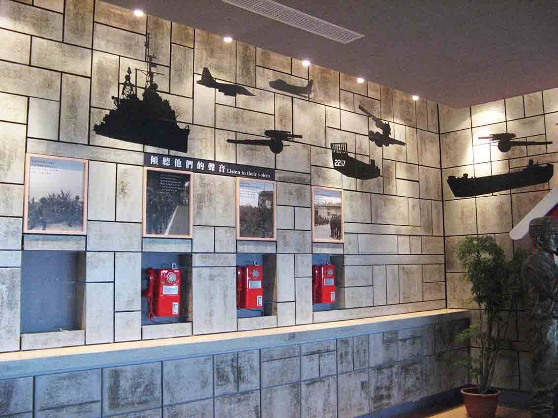 湖井頭戰史館「傾聽他們的聲音」展牆，水泥磚紋背景搭配軍艦戰機剪影與紅色野戰電話展示