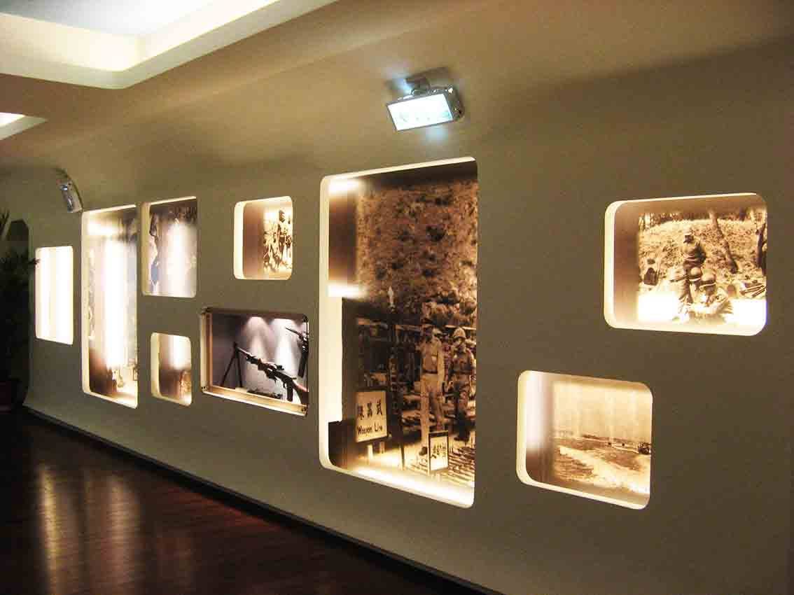 湖井頭戰史館暖光展廊全景，多幅黑白戰地舊照與輕兵器壓克力展示窗沿牆排列，呈現古寧頭戰役歷史氛圍